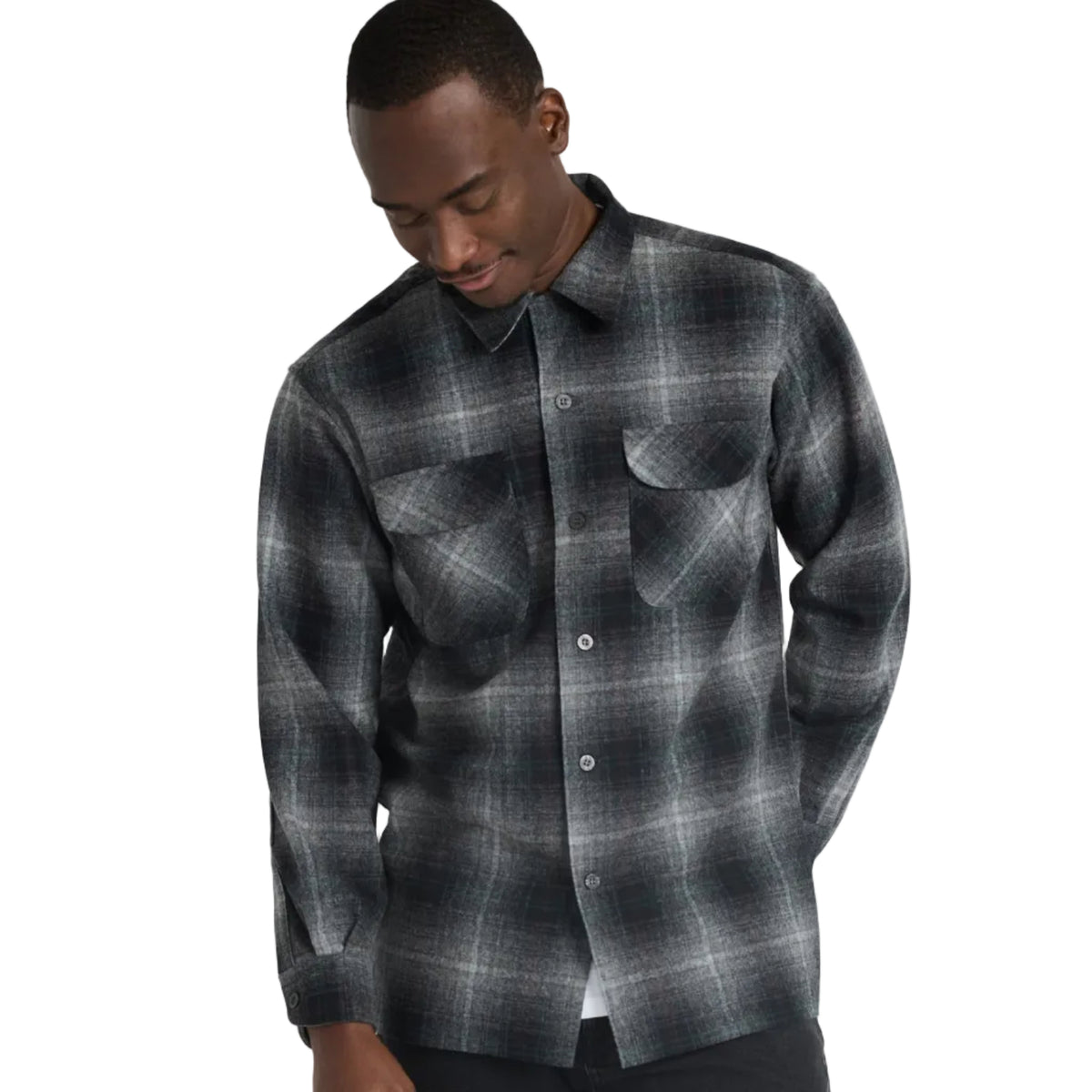 A man wearing a dark gray and black plaid button-down shirt with two chest pockets, paired with black pants, photographed against a light gray studio background.