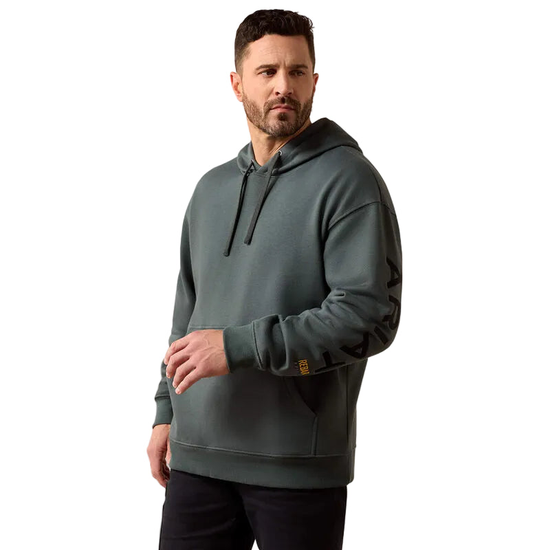 A man wearing a dark slate green pullover hoodie with black drawstrings and a black graphic print on the left sleeve, standing against a plain beige background.