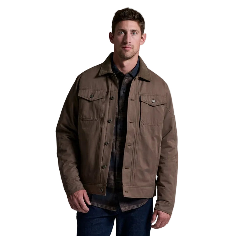 A man wearing a brown button-up field jacket over a dark shirt and blue jeans, standing against a solid gray studio background.